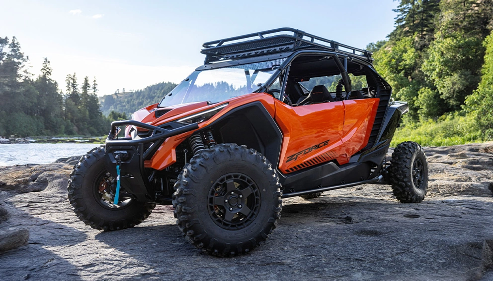 CFMoto ZForce orange équipé d'un porte-bagages de toit, garé sur des rochers au bord d'une rivière.