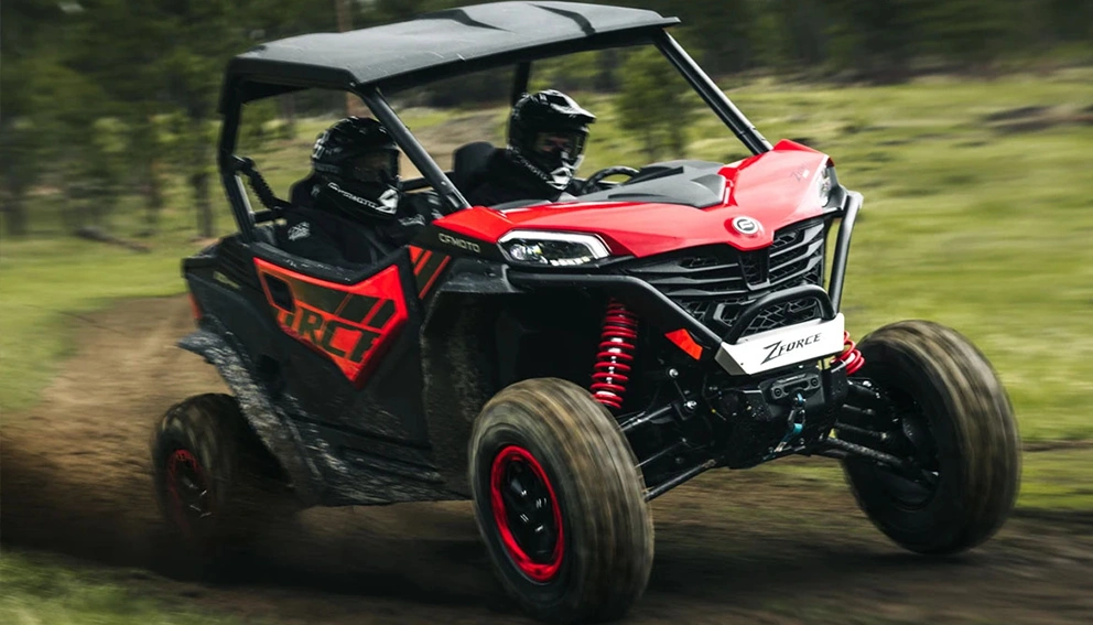 CFMoto ZForce 950 Sport rouge et noir en pleine action, deux passagers, projetant de la terre sur un chemin herbeux.