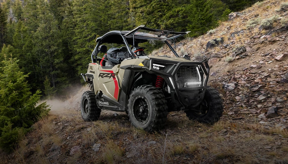 Polaris RZR Trail Ultimate 2026 beige gravissant un sentier rocailleux en montagne, forêt en arrière-plan.