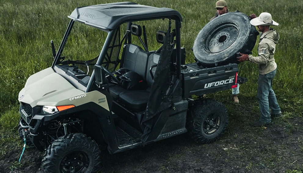 CFMoto UForce 600 2026 beige garé dans un champ, deux personnes chargeant un pneu à l'arrière.