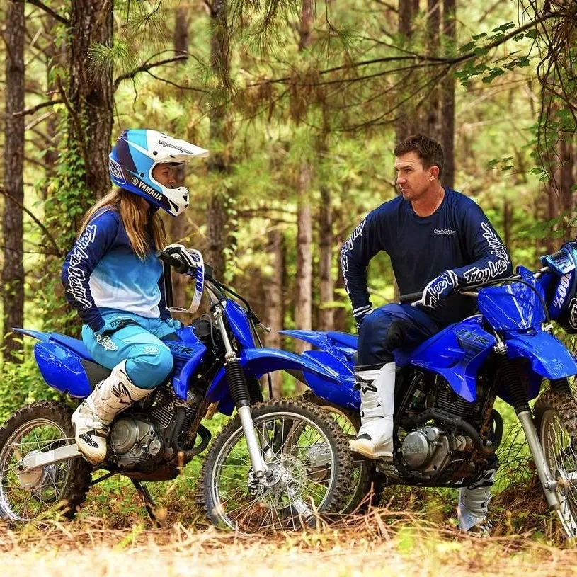 Jeune pilote sur une Yamaha TT-R 125 bleue accompagné d'un adulte en forêt.