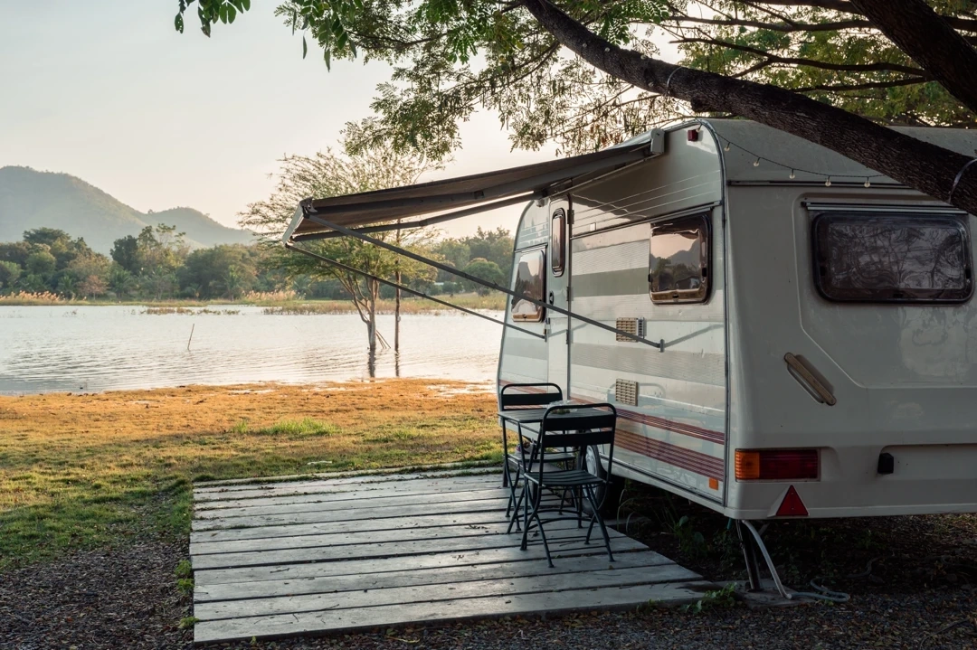 Roulotte stationnée au bord d’un lac avec un auvent déployé, deux chaises et une table installées sur une terrasse en bois, ambiance paisible au coucher du soleil.