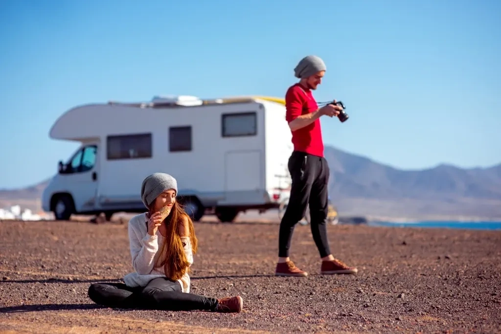 Enfant assise au sol et adulte prenant des photos près d’un VR stationné, représentant un voyage en roulotte flexible et adapté aux familles.