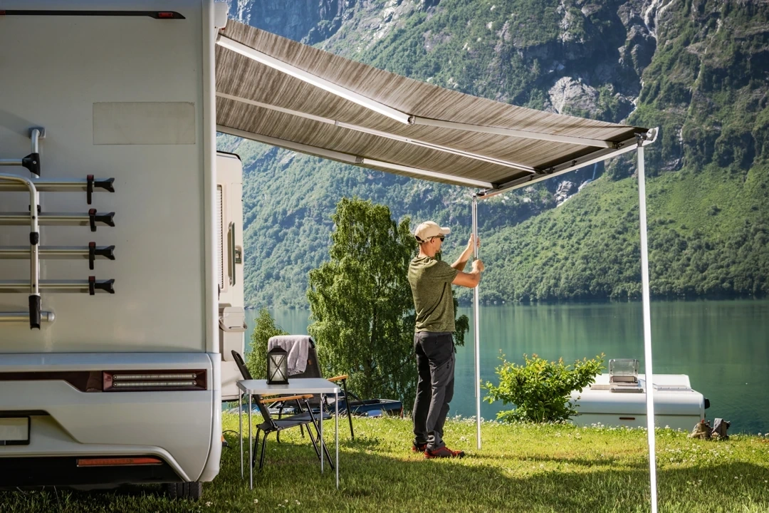 Camping-car garé en bord de lac avec un auvent déployé, homme ajustant un poteau d’auvent face à un paysage de montagnes verdoyantes et d’eau calme.