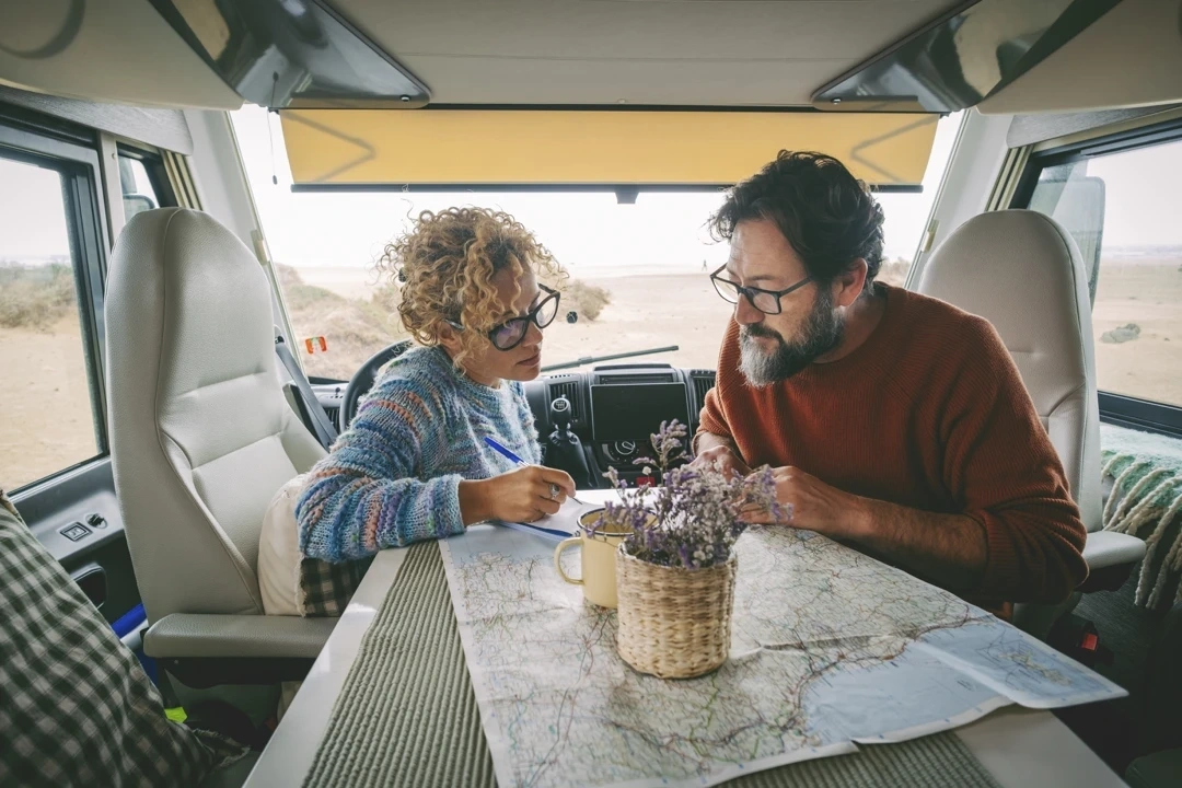 Couple assis dans un VR stationné au bord d’une plage, concentrés sur une carte routière étalée sur la table, ambiance calme avec lumière naturelle à travers les fenêtres.
