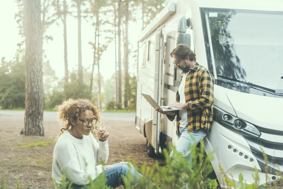 Scène matinale dans un camping en Gaspésie : femme assise au sol parmi les plantes, homme debout avec un ordinateur portable près d’un véhicule récréatif.