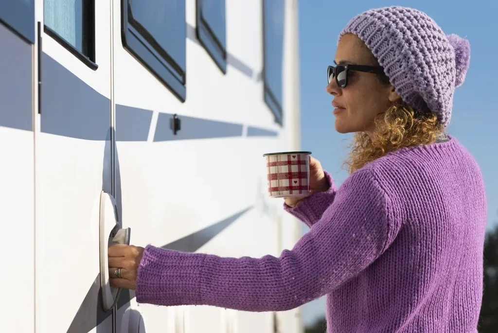 Femme ouvrant la porte d’une roulotte en hiver avec une boisson chaude, représentant l’inspection et l’achat d’une roulotte durant la saison hivernale.