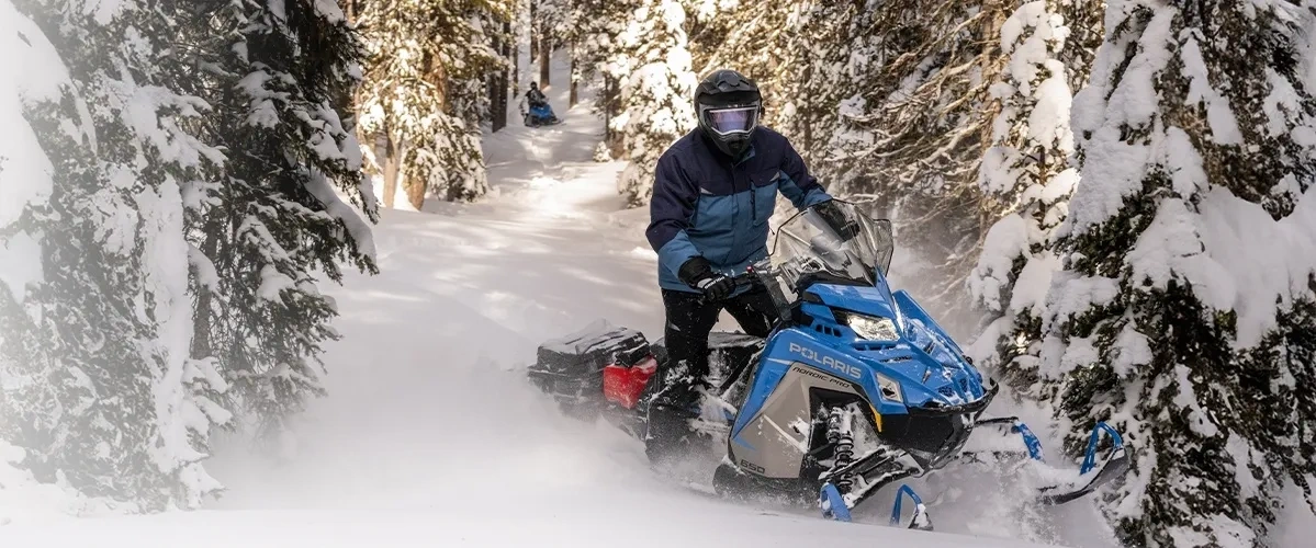 Motoneigiste au guidon d’une Polaris bleue sur un étroit sentier bordé d’arbres enneigés.