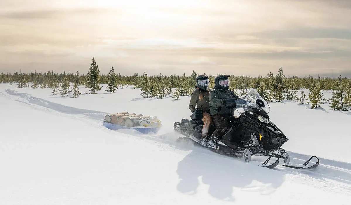 Polaris Voyageur Adventure Ultimate 2027 avec deux occupants remorquant un traîneau chargé dans la neige.