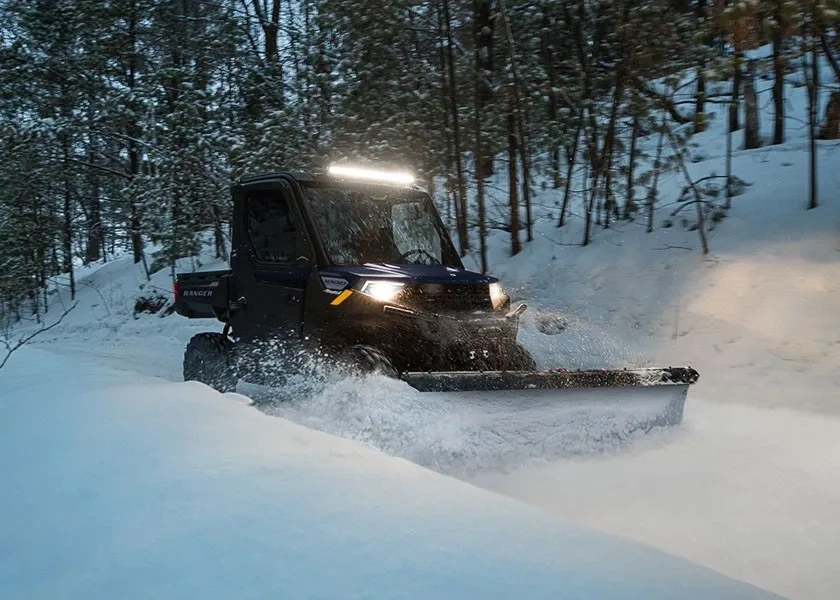 Polaris RANGER 1000 CAB 2027 équipé d’une lame de déneigement sur un chemin forestier enneigé.