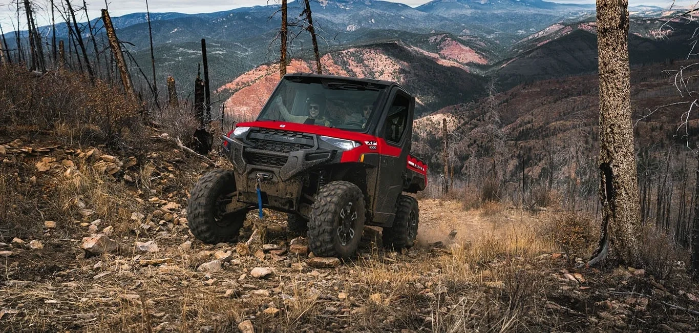 Polaris RANGER XP 1000 CAB rouge sur un terrain rocailleux avec des montagnes à l’arrière-plan.
