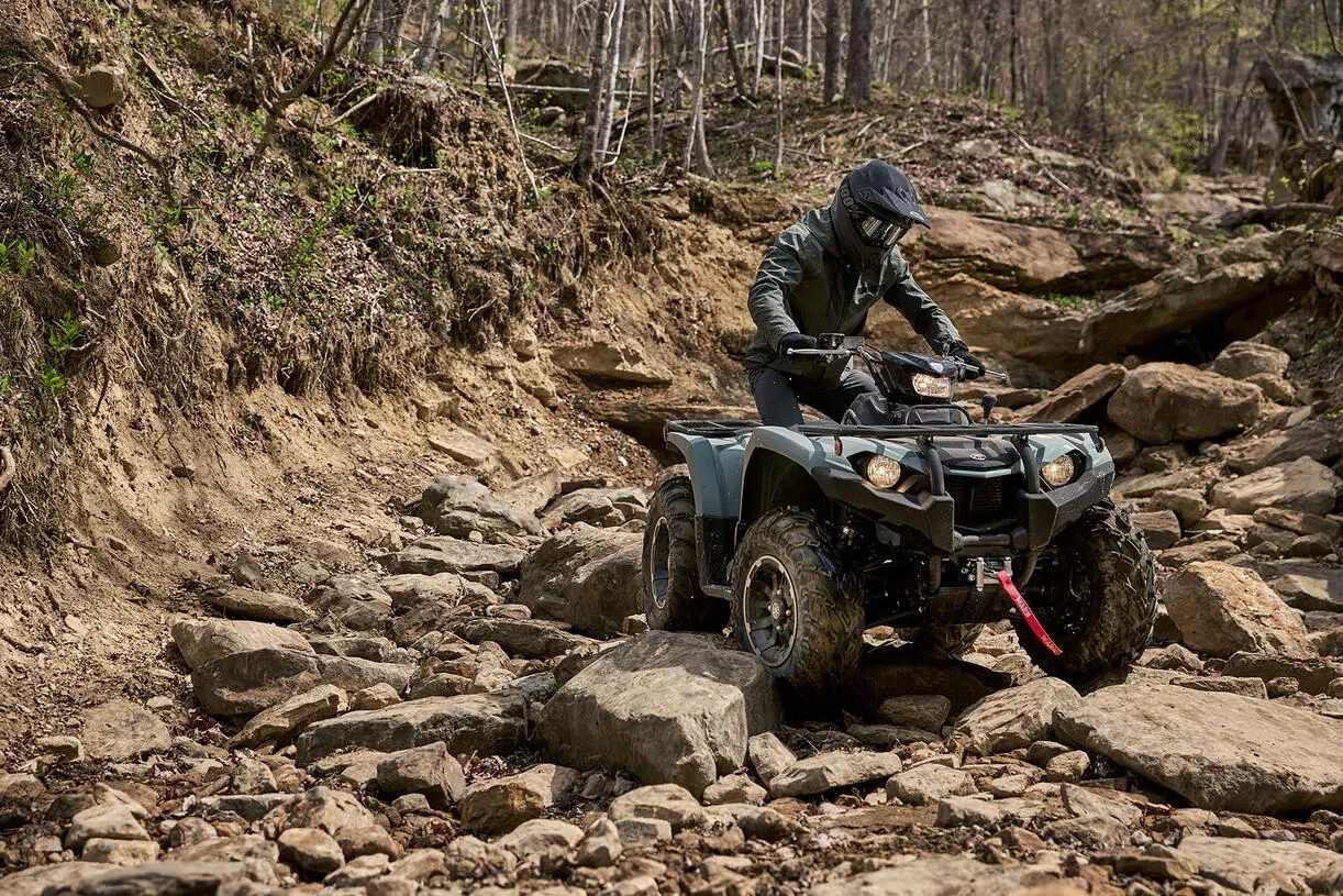 Pilote sur un VTT Yamaha Kodiak 450 franchissant un sentier rocheux en forêt.