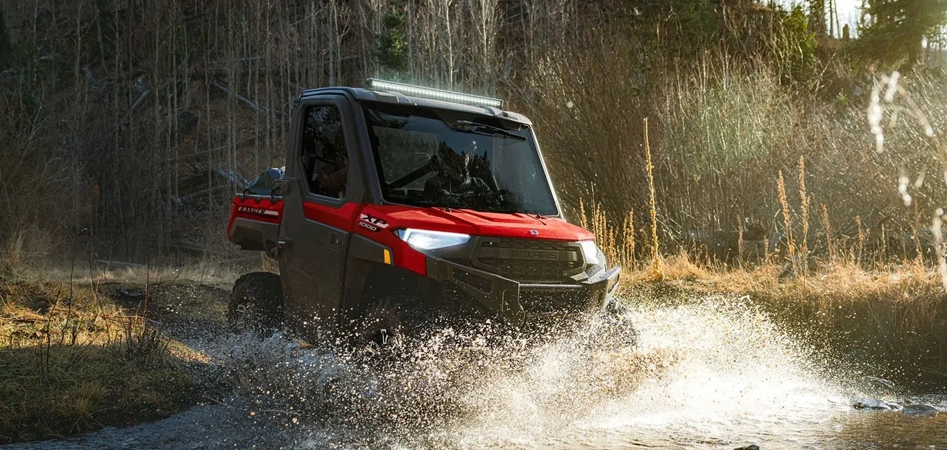 Polaris RANGER XP 1000 CAB rouge traversant une flaque d’eau sur un chemin forestier.