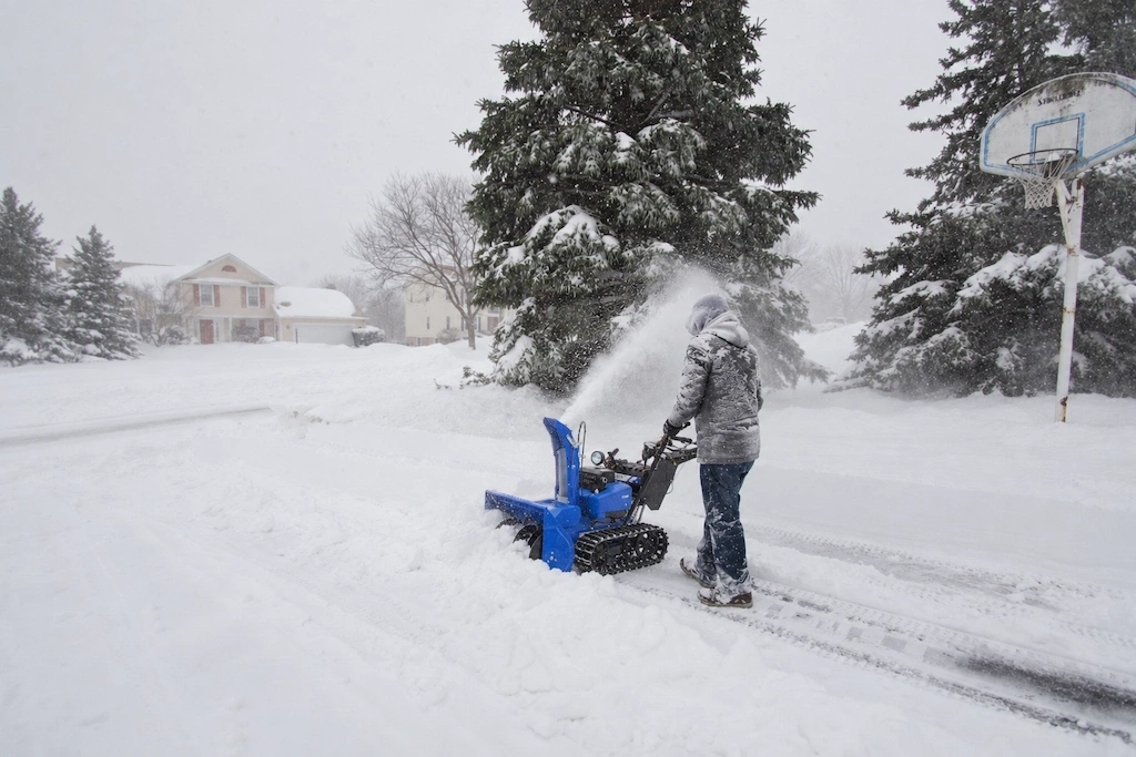 Person clearing a residential driveway with a blue snowblower during snowfall.