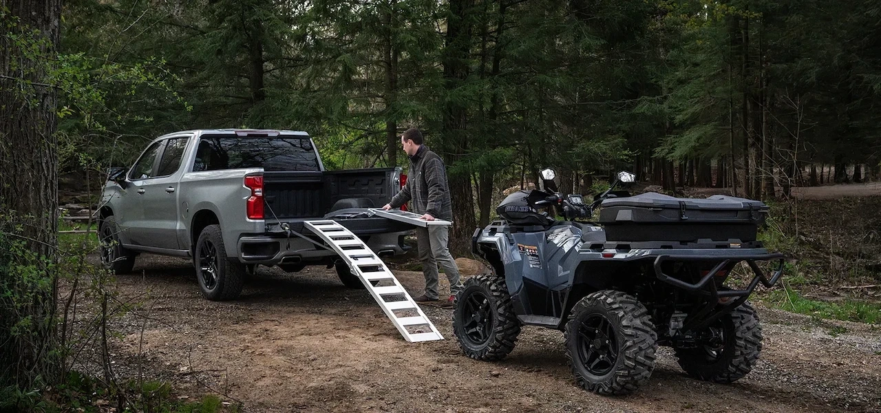 VTT Polaris Sportsman 500 2027 équipé d’un coffre arrière près d’une camionnette avec des rampes de chargement en forêt.