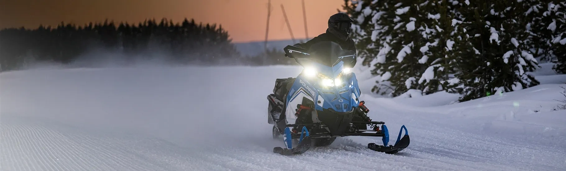 Motoneige Polaris INDY VR1 9R 2027 bleue avec ses phares allumés sur un sentier enneigé au crépuscule.