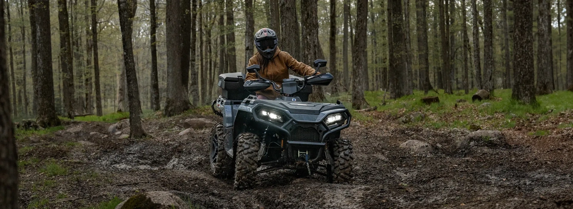 Polaris Sportsman 500 2027 avec ses phares allumés sur un sentier boueux en forêt.