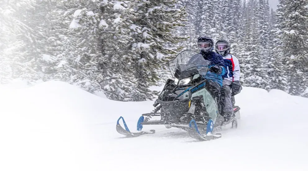 Motoneige Polaris Indy Adventure 2026 bleue transportant deux passagers à travers une tempête de neige en montagne.