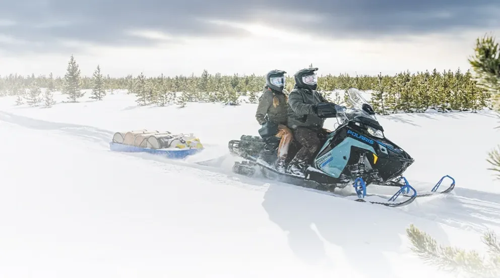 Motoneige Polaris Voyageur Adventure Ultimate 155 2026 bleue avec deux passagers traversant une piste enneigée.